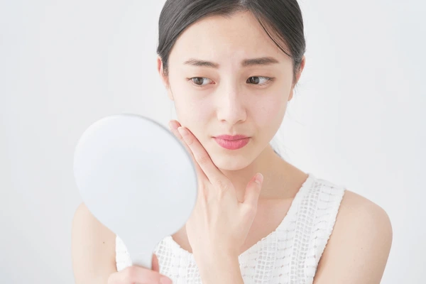 Concerned woman examining her skin in a mirror, representing potential side effects and risks associated with Dysport treatment.